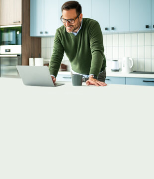 Man smiling and focusing on his laptop while standing at a counter in a modern kitchen, managing business tasks, legal work, or accounting from a comfortable remote location