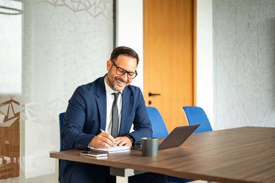 Professional man in a business suit working at a conference table. Actively writing notes in a notebook while using a laptop and a mobile device