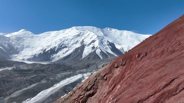 Aerial video pan revealing snow-capped Lenin Peak behind a steep red mountain slope under a clear blue sky