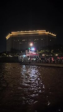 Vertical night view of a vehicle ferry moored and crowded with passengers and cars,  with its reflection on the windy Mekong River and a large illuminated hotel in the background.