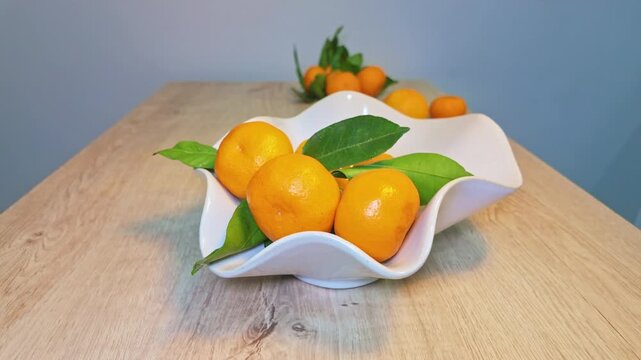 Fresh ripe mandarins with green leaves arranged in a white wavy ceramic bowl on wooden table.