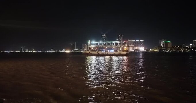 Night view of Phnom Penh skyline featuring a cruise boat on the Mekong River with searchlight beams from a skyscraper in the background.