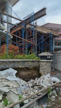 Blue metal scaffolding surrounds an unfinished building structure with exposed steel beams, while broken tiles and bamboo poles lie scattered on the ground below a weathered stone wall.