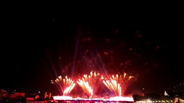 Panoramic night view of Golden and red firework explosion display shooting upward from a bridge over riverfront city background during a festival in dark night time sky.