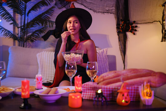 Young Asian woman in a witch costume enjoying snacks at a Halloween party table.