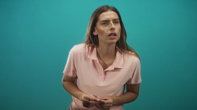Woman in a studio wearing a pink polo, hands fidgeting and fingers clasped, mouth parted and leaning forward against teal backdrop; confusion.