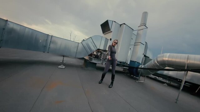 Young woman striking expressive dance pose on industrial rooftop beside silver ducts under gray sky