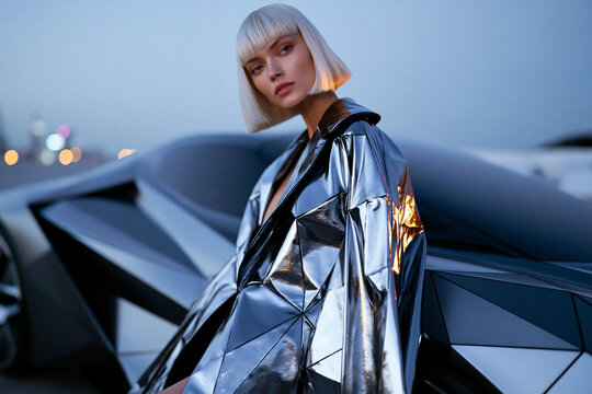 Blonde Frau in silbernem Metallic-Outfit lehnt an futuristischem Auto