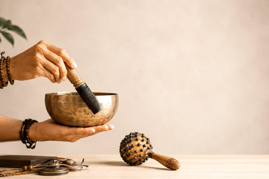 Peaceful meditation background template with ample negative copy space, featuring hands playing a bronze singing bowl with a wooden striker, beads, and a rattle on a wooden table.