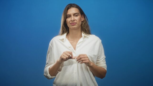 Woman pressing fingertips together, fidgeting with white shirt button against blue studio backdrop; unease hesitation.