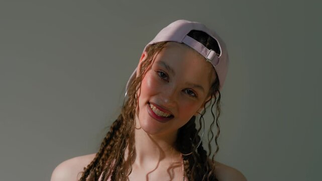 Smiling young woman with backward pink cap, braids, hoop earrings, sunlight against green background