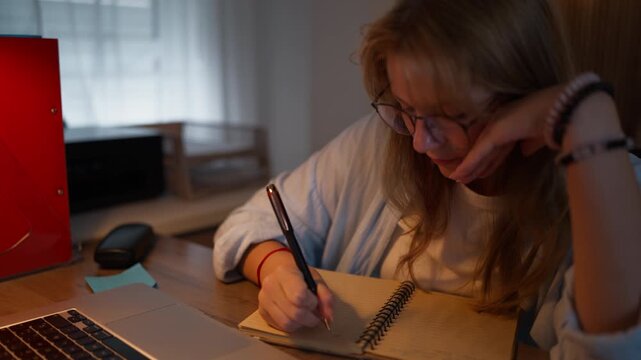 Focused teenage student wearing glasses taking handwritten notes in a notebook with a laptop beside her. Note-taking, study skills, learning, concentration and academic development concept.