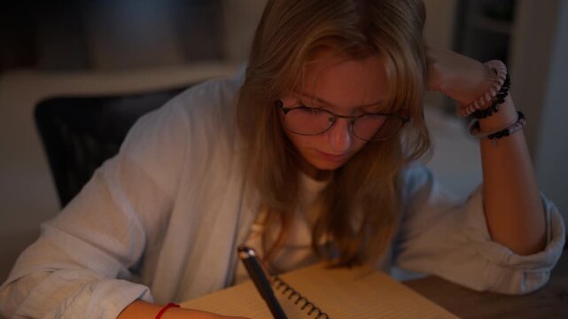 Teenage girl wearing eyeglasses taking handwritten notes beside a laptop during an evening study session. Blended learning, digital education, schoolwork and modern study habits concept.