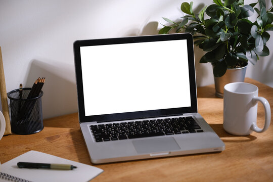 Modern home office workspace with blank laptop screen, notebook, headphones, clock and stationery arranged on a wooden desk with natural light.