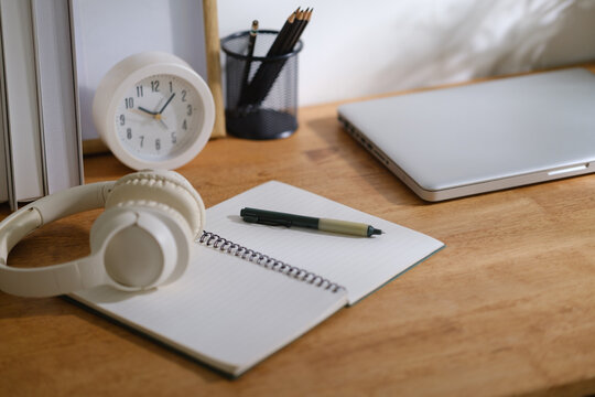 Minimal home office desk with notebook, pen, headphones, clock and closed laptop on wooden table in soft natural light.