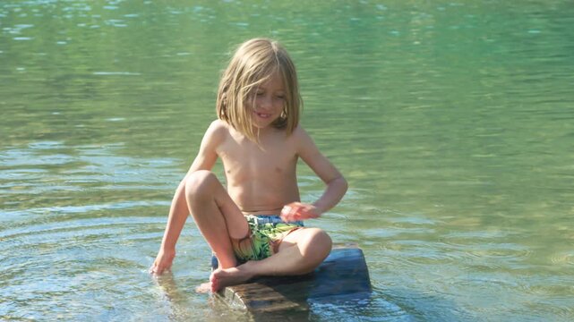 Boy laughing joyfully as water splashes over him while sitting on a river rock