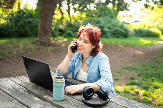 Smiling businesswoman talking on phone and using laptop at outdoor park office.
