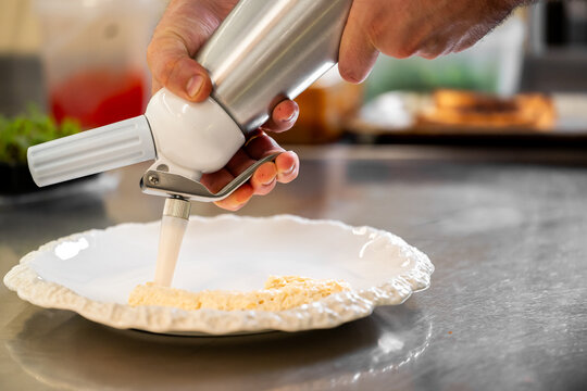 A chef uses a professional whipped cream dispenser to pipe a light, creamy espuma or mousse onto a decorative white plate in a commercial kitchen setting