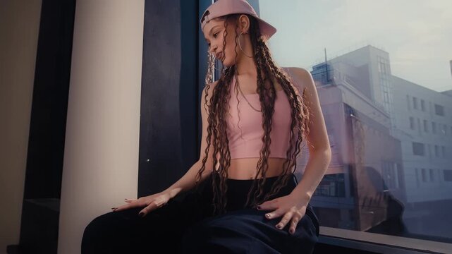 Confident young woman in pink cap posing beside glass building under sunny daylight urban style pose
