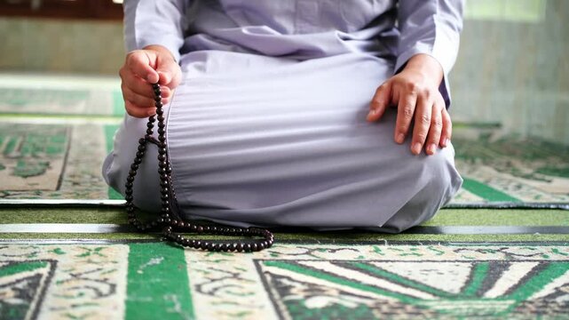 A close-up photo of a hand performing dhikr with prayer beads inside a mosque. A hand engaged in dhikr. A man performing dhikr while holding prayer beads. Front-facing shot.
