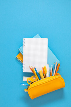 Vertical flat lay of school stationery featuring an open yellow pencil case filled with colorful pens and pencils below a blank grid spiral notebook on a vibrant blue background with copy space.
