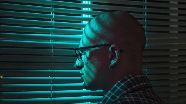 Caucasian young adult man looking through window blinds with blue light reflecting on face. Clip is ideal for mental health campaigns