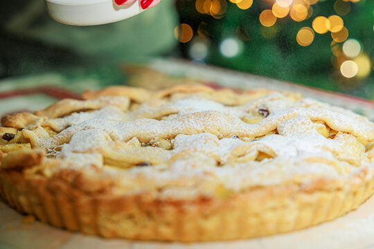 dettagli di una crostata alla frutta fatta in casa mentre viene ricoperta da dello zucchero a velo, sfondo sfuocato con luci e decorazioni natalizie