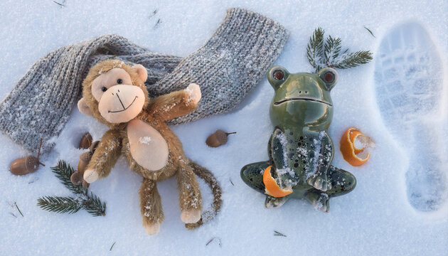Playful monkey plush and a ceramic frog resting on a fresh winter snow background with scarf and pinecones