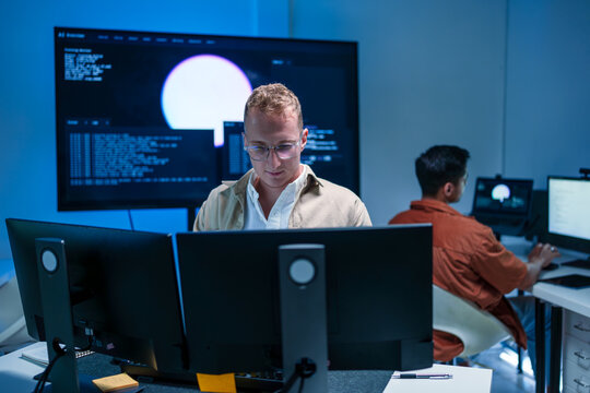 Young male software developer working on code and data analysis with multiple monitors in tech office laboratory