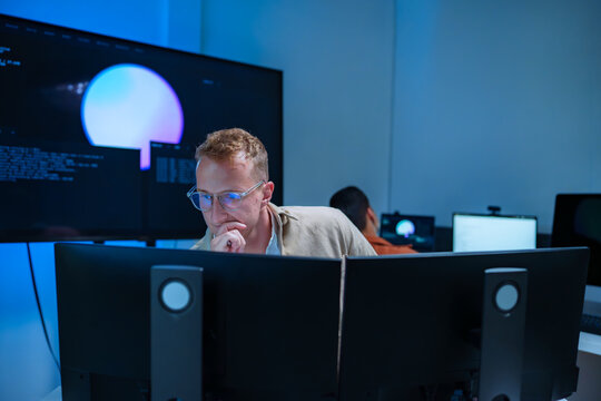 Young male developer in clear glasses deeply focused on multiple computer monitors writing code and analyzing AI system workflows in a modern blue lit tech office