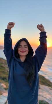 Young woman celebrating at sunset on coastal cliff in hoodie for lifestyle background and wellness poster design