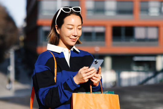 Young asian woman smiling looking at her smartphone and holding shopping bags in an urban environment on a sunny day