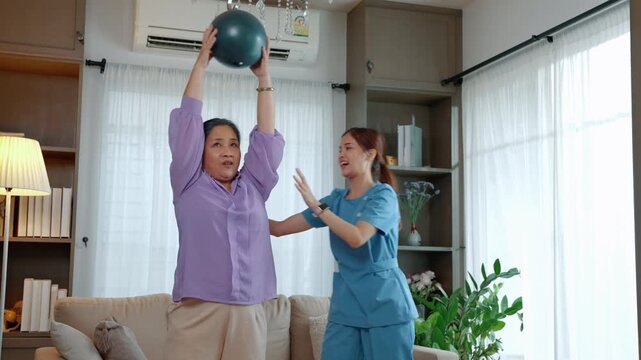Senior woman exercising with a caregiver using a fitness ball at home, representing rehabilitation, physical activity, healthy aging, mobility, and wellness.
