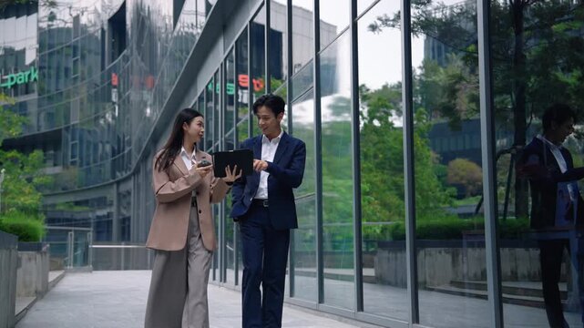 Two business professionals walking through a modern office complex while reviewing information on a tablet, representing teamwork, collaboration, technology, and corporate work.
