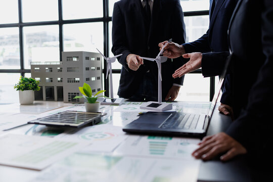 Business people collaborating on sustainable development, analyzing models of wind turbines, a solar panel, and a building on a desk