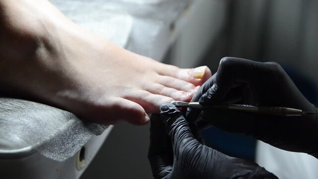 Pedicurist gently pushes back the cuticle on a woman's toenails using a metal cuticle pusher. Close-up professional pedicure and nail preparation procedure.