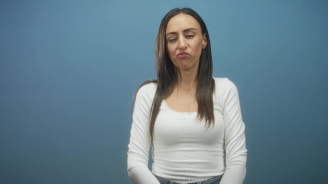 Young woman in white top purses her lips and checks a smartphone with one hand while seated against a blue backdrop in a studio; annoyance.