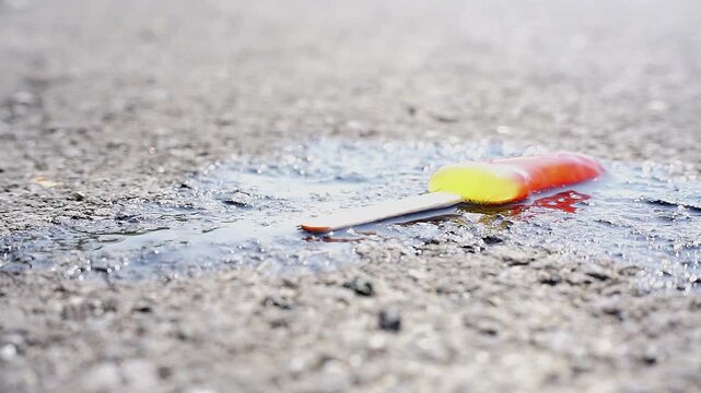 Melted colorful popsicle on a wooden stick on rough asphalt under bright sunlight