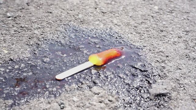Melted colorful popsicle on a wooden stick on rough asphalt under bright sunlight