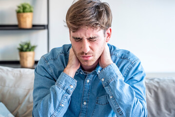 Fototapeta premium Young man with neck pain and stress sitting on couch holding his neck in visible discomfort