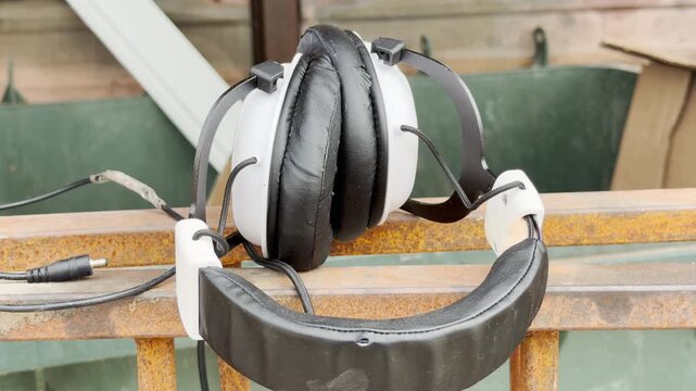 White and black headphones rest on a rusty metal rail outdoors. The discarded headset hangs quietly in daylight. The abandoned object evokes a lonely, nostalgic feel.