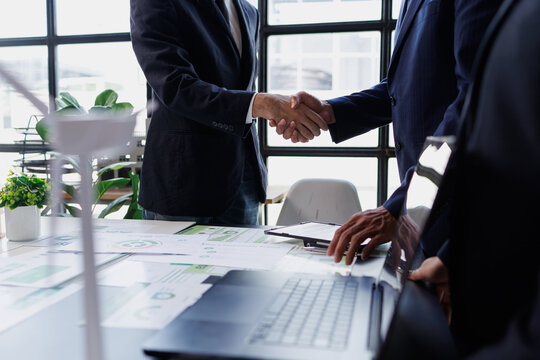 Business partners shaking hands over desk with green energy charts, confirming sustainable deal