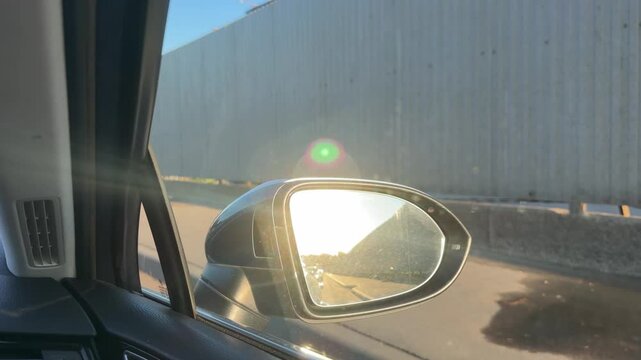Car side mirror captures sunlight and reflections of industrial buildings while parked along a road next to a metal fence under a blue sky