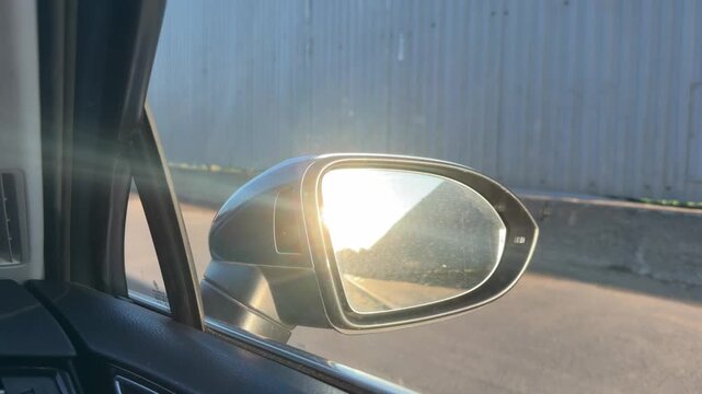 Side view mirror reflects sunlight as the car moves along a road next to a corrugated metal wall under a clear blue sky