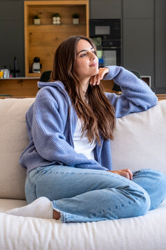 Young woman relaxing on a sofa at home, wearing cozy sweater and jeans, looking thoughtful and peaceful in a modern living room setting.