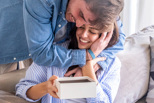 Man hugging smiling woman as she opens a gift box at home, romantic surprise between happy couple sharing affection and joy on couch