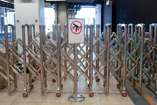 Barriers stand arranged in a crisscross pattern, signaling caution in a transit hub. Sunlight filters through the glass, casting dynamic shadows on the floor