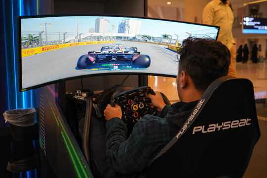 A young man playing an advanced Formula 1 racing simulator game with a curved monitor screen, racing steering wheel, and cockpit rig set up at an interactive indoor venue in Jeddah, Saudi Arabia.