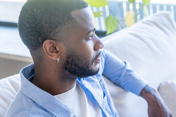 Young bearded man sitting on a sofa looking thoughtful in profile at home, casual shirt and earring conveying relaxed lifestyle and quiet contemplation © simona