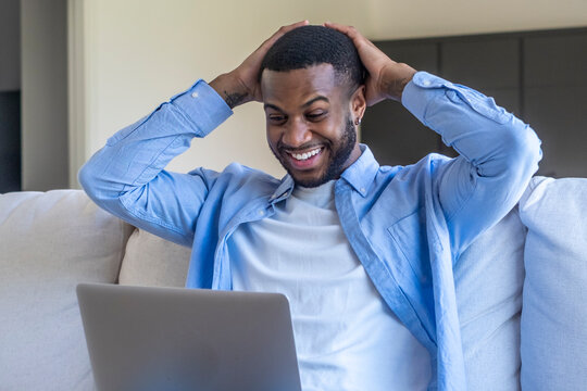 Smiling man relaxing on a couch with a laptop, celebrating good news while working from home and enjoying remote work, happiness and casual productivity